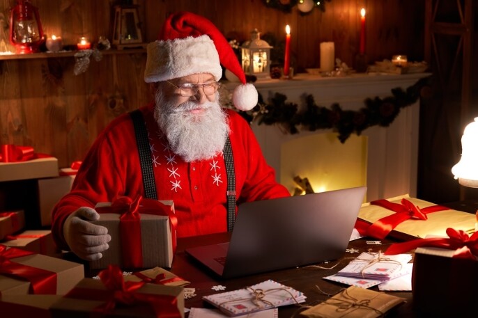 Happy old Santa Claus wearing hat holding gift box using laptop computer sitting at workshop home table late on Merry Christmas eve. Ecommerce website xmas time holiday online shopping e commerce sale